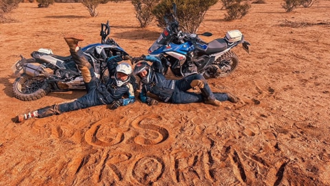 Deux pilotes de l’équipe féminine internationale sont allongées devant leur moto dans le sable où elles ont écrit « GS Trophy ».